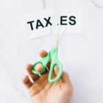 Vue de dessus d'une main tenant des ciseaux coupant le mot « taxes » sur une surface en marbre.