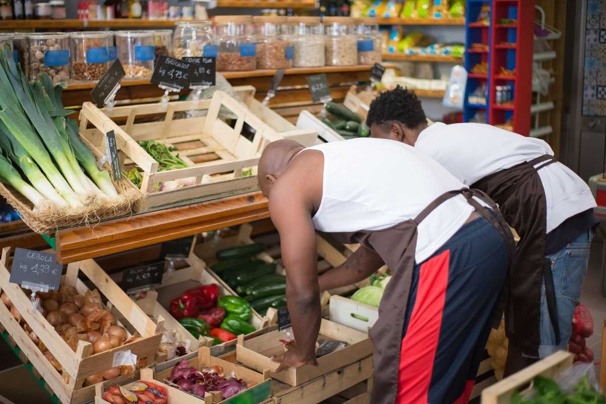 Homme en blouse blanche et tablier, manipulant des légumes dans un magasin