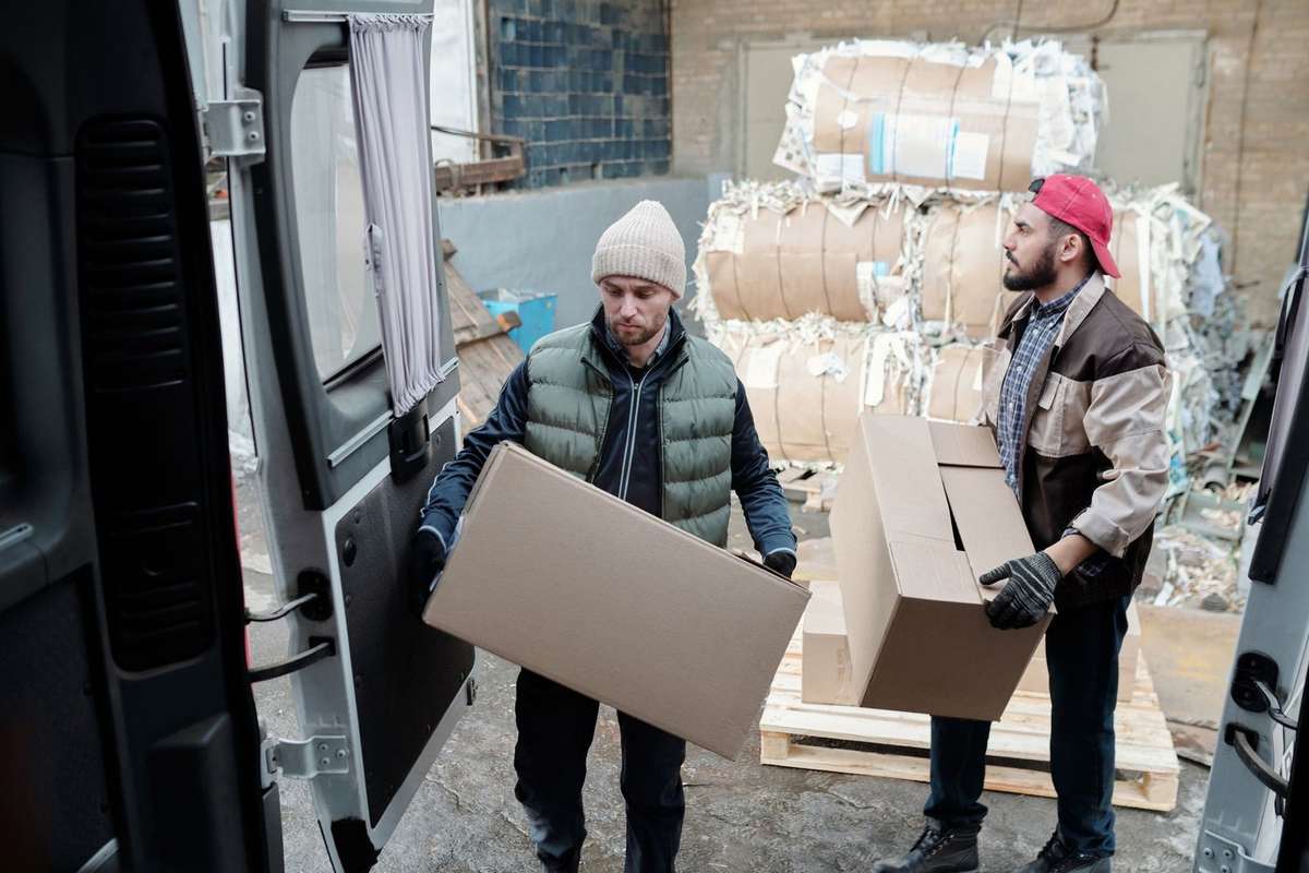Homme en vestre décharge une boîte, autre en rouge décharge une autre, camion ouvert, entrepôt en arrière-plan